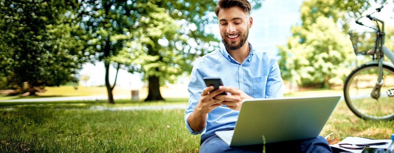 Man sitting on a grassy field in a park next to a parked bike. He is smiling and looking at a phone in his hands while a laptop is sitting on his lap.